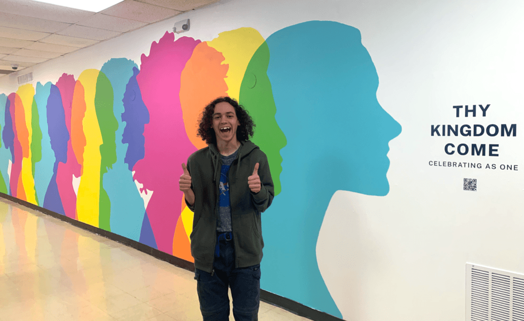 Student Korey Rees smiling and giving two thumbs up in front of the Thy Kingdom Come Mural